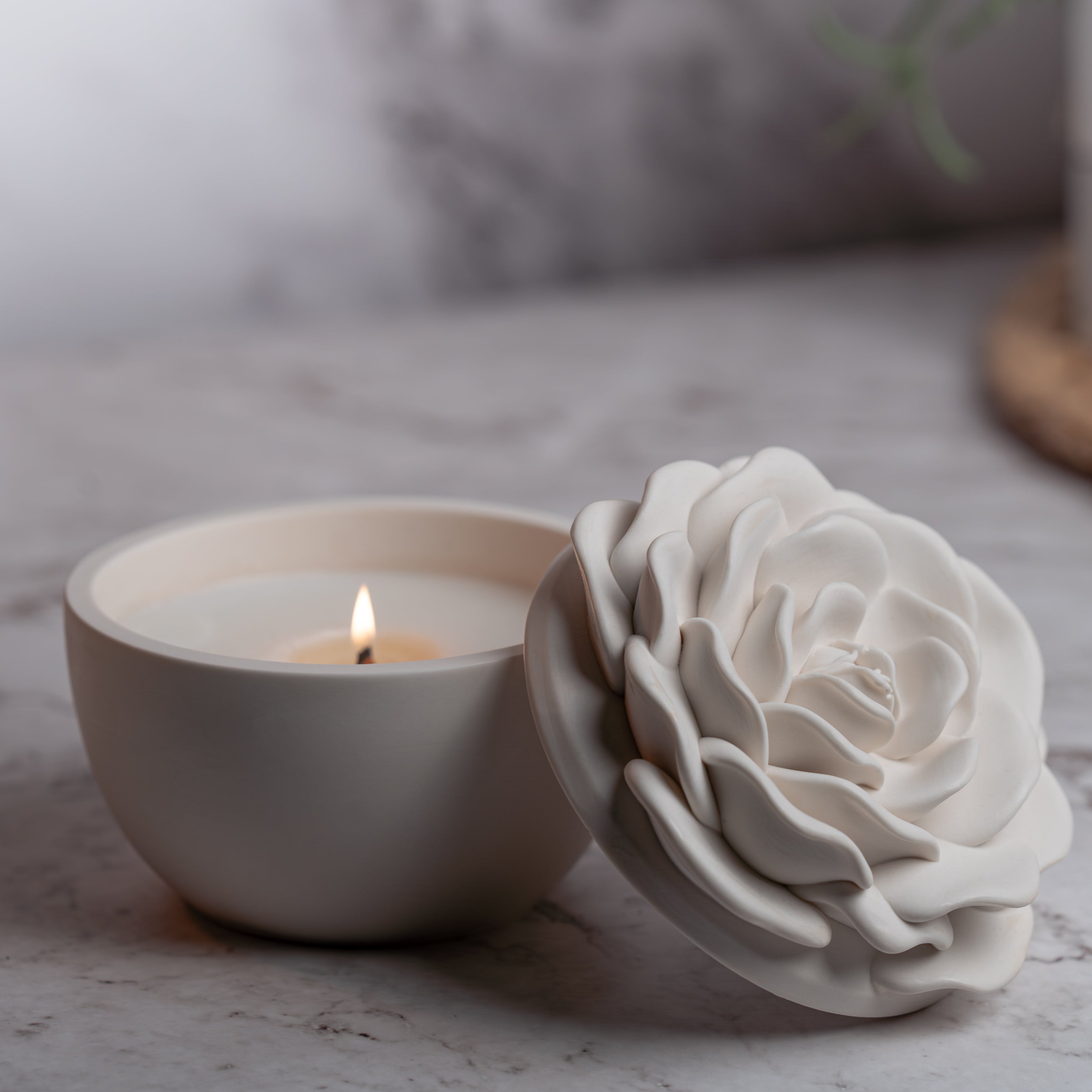 White ceramic rose next to a lit candle in a bowl on a marble surface.